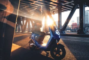 Le Niu MQi+ Sport bleu sous un pont au soleil couchant