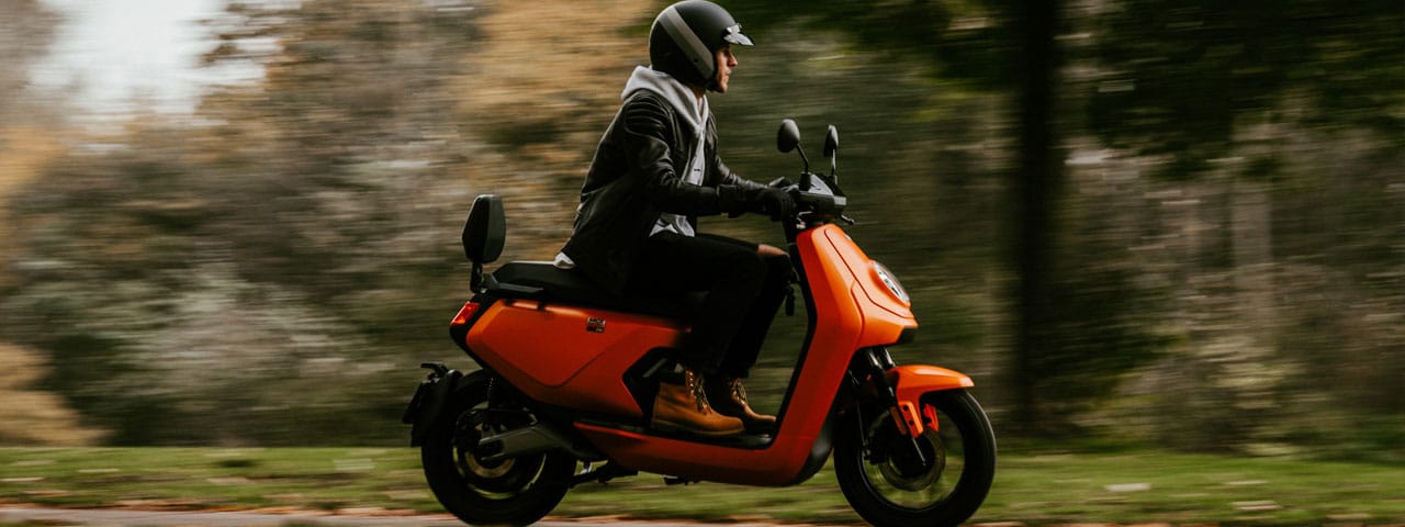 Niu MQi GT orange et son conducteur à vive allure sur une route de campagne